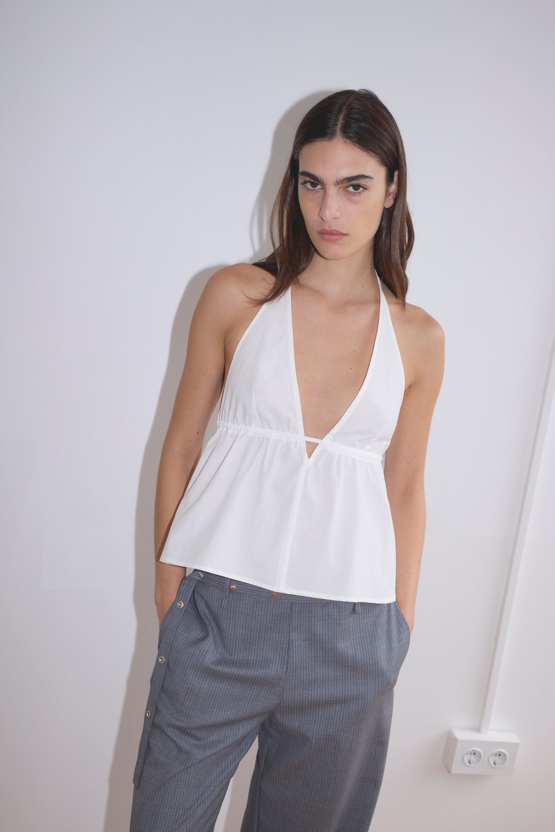 A person with long brown hair wears the Button Halter - White, a sleeveless organic cotton halter top, paired with loose gray pinstripe pants, standing against a plain white wall with hands in pockets.