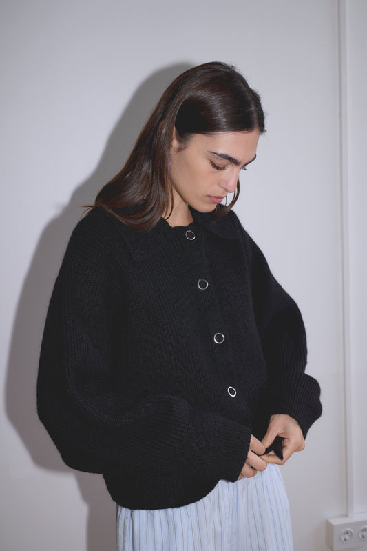 A woman with long brown hair buttons the Stud Cardi Knit - Black, a sustainable oversized cardigan, while standing against a white wall and wearing light blue striped pants.
