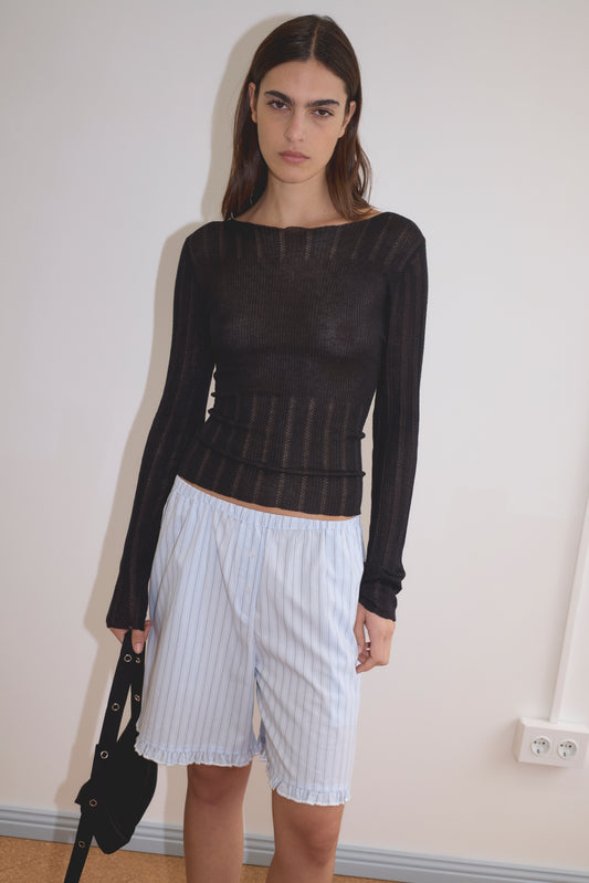 A woman stands indoors against a plain wall, wearing the Open Work Longsleeve in black by Deiji Studios and loose, light blue pinstriped shorts. She holds a black bag and looks directly at the camera with a neutral expression.