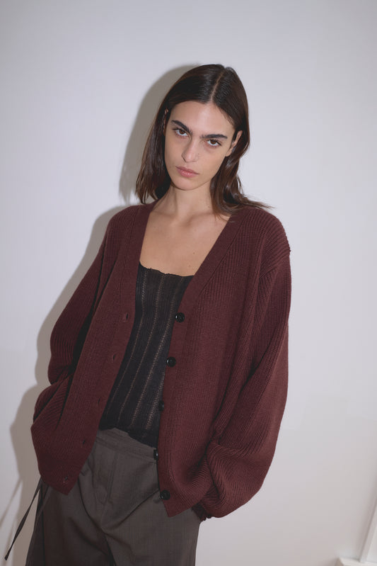 A woman with straight brown hair wears the Flounce Cardigan - Burgundy by Deiji Studios over a black top and brown pants, standing against a plain white background with a neutral expression.