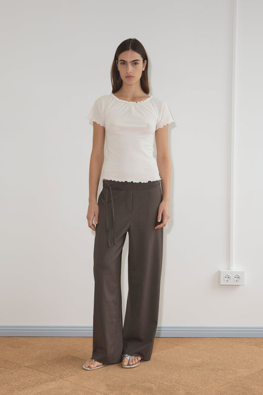A woman stands indoors against a white wall, wearing a short-sleeved organic cotton white top, Double Belted Pant - Brown Stripe, and silver sandals. The floor is light brown, with two power outlets visible on the wall.