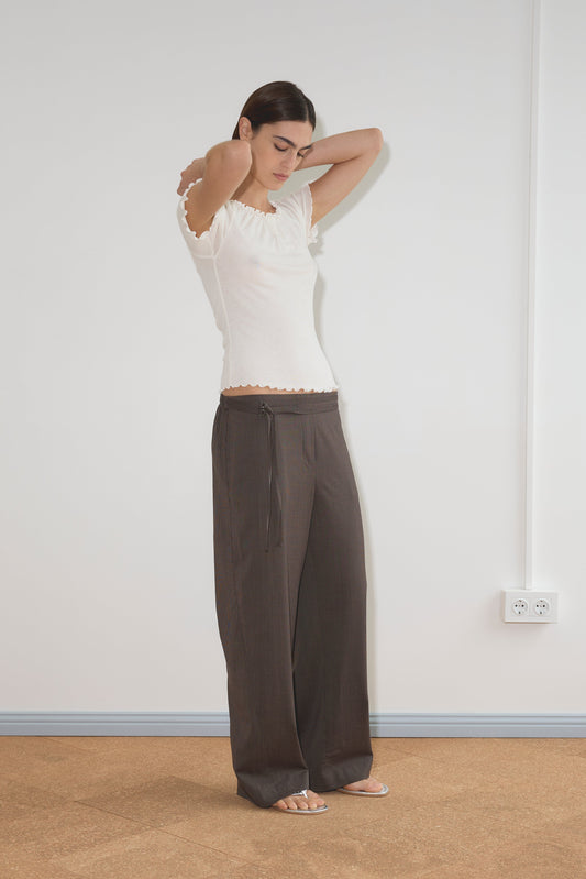 A woman stands indoors against a plain wall, wearing a white short-sleeve top and the Double Belted Pant - Brown Stripe. She has her arms raised, adjusting her hair, and is wearing sandals.