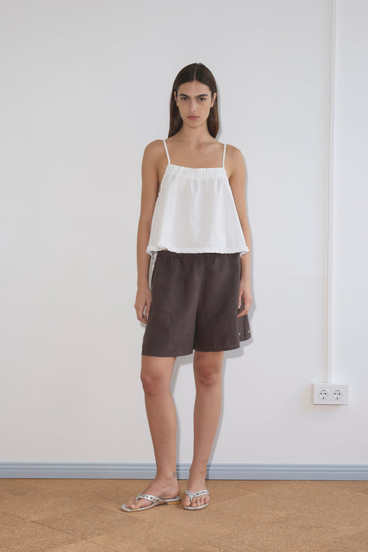 A woman with straight brown hair and a neutral expression stands indoors against a white wall, wearing the Yoke Top - White by Deiji Studios in organic cotton poplin, paired with brown knee-length shorts and white sandals.