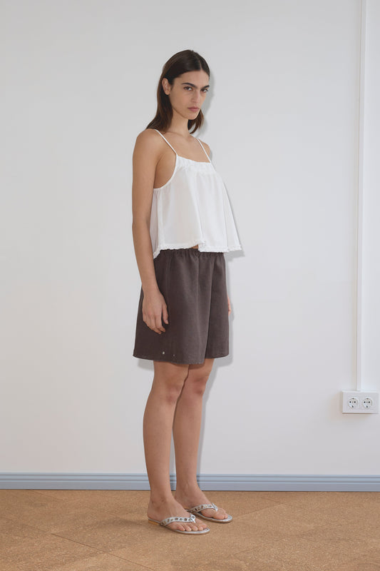 A woman with straight brown hair and a neutral expression stands indoors against a plain white wall, wearing the Yoke Top - White by Deiji Studios, paired with brown shorts and clear flat sandals.