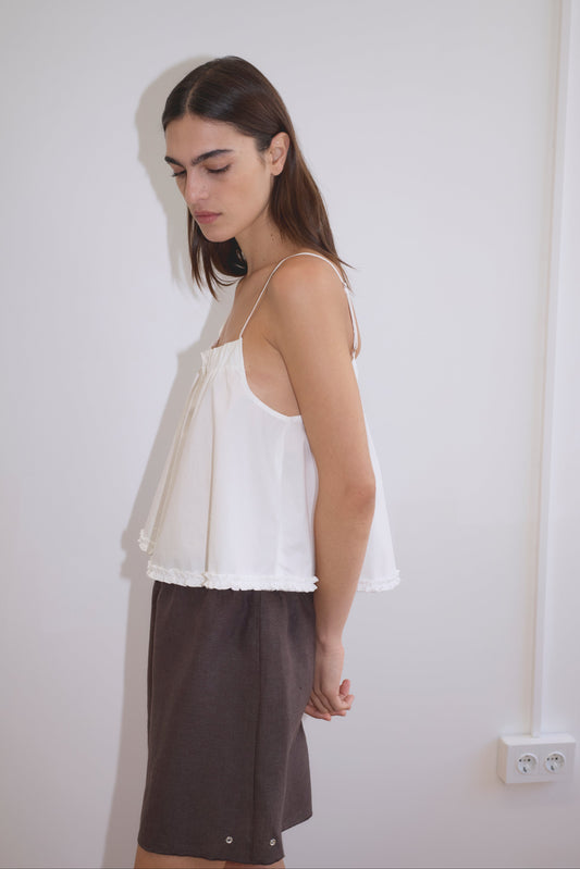 A woman with long brown hair stands sideways against a plain white wall, wearing a loose white sleeveless top from Deiji Studios and the Press Stud Short - Bark. Her head is slightly bowed and her hands are clasped behind her back.