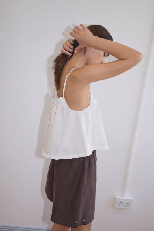 Wearing the Yoke Top - White, a person in dark shorts stands sideways against a white wall, lifting their hair with both hands. Their face is partially turned away, reflecting the relaxed style of Deiji Studios.