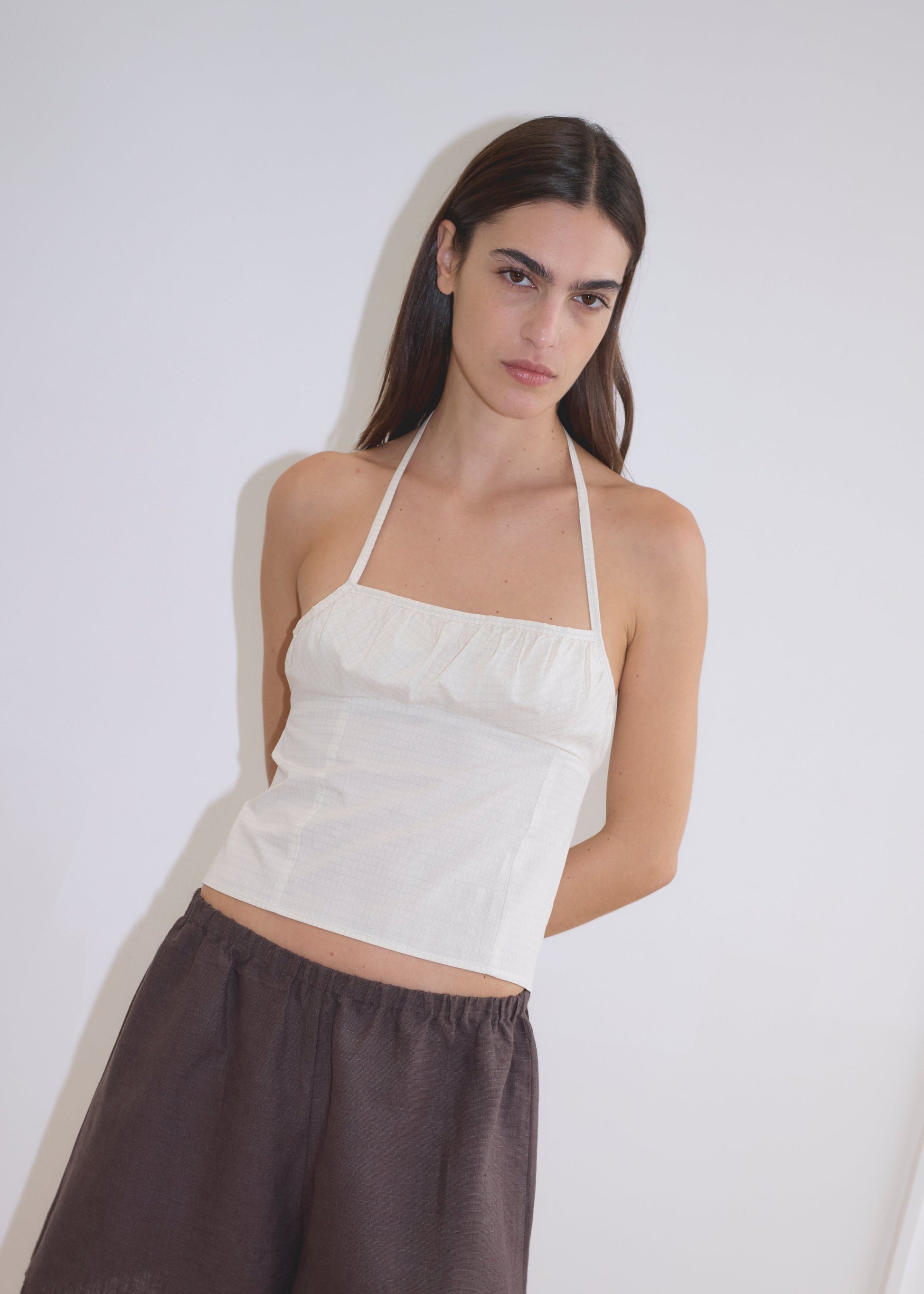 A woman with long dark hair wears a white Strap Top and loose brown shorts, standing against a plain white background with her hands behind her back.