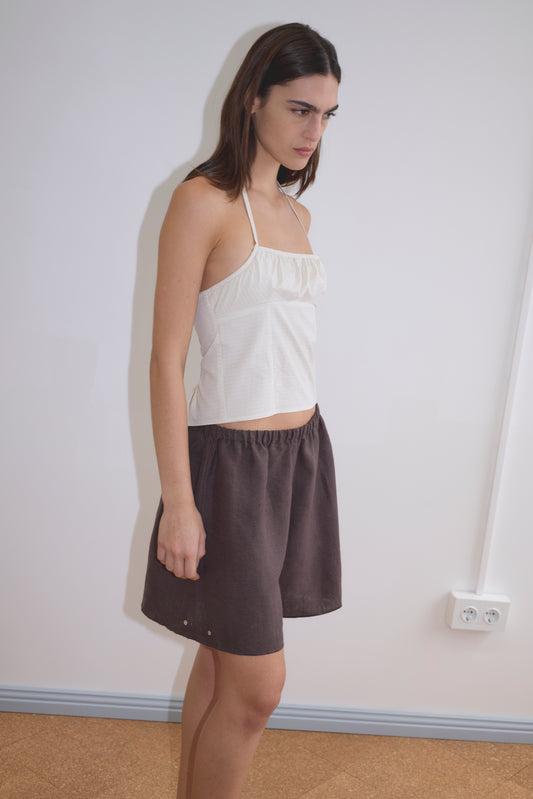 A woman stands indoors against a white wall, wearing the Strap Top - Bone Check with thin straps and loose dark brown shorts. She has straight brown hair and a neutral expression.