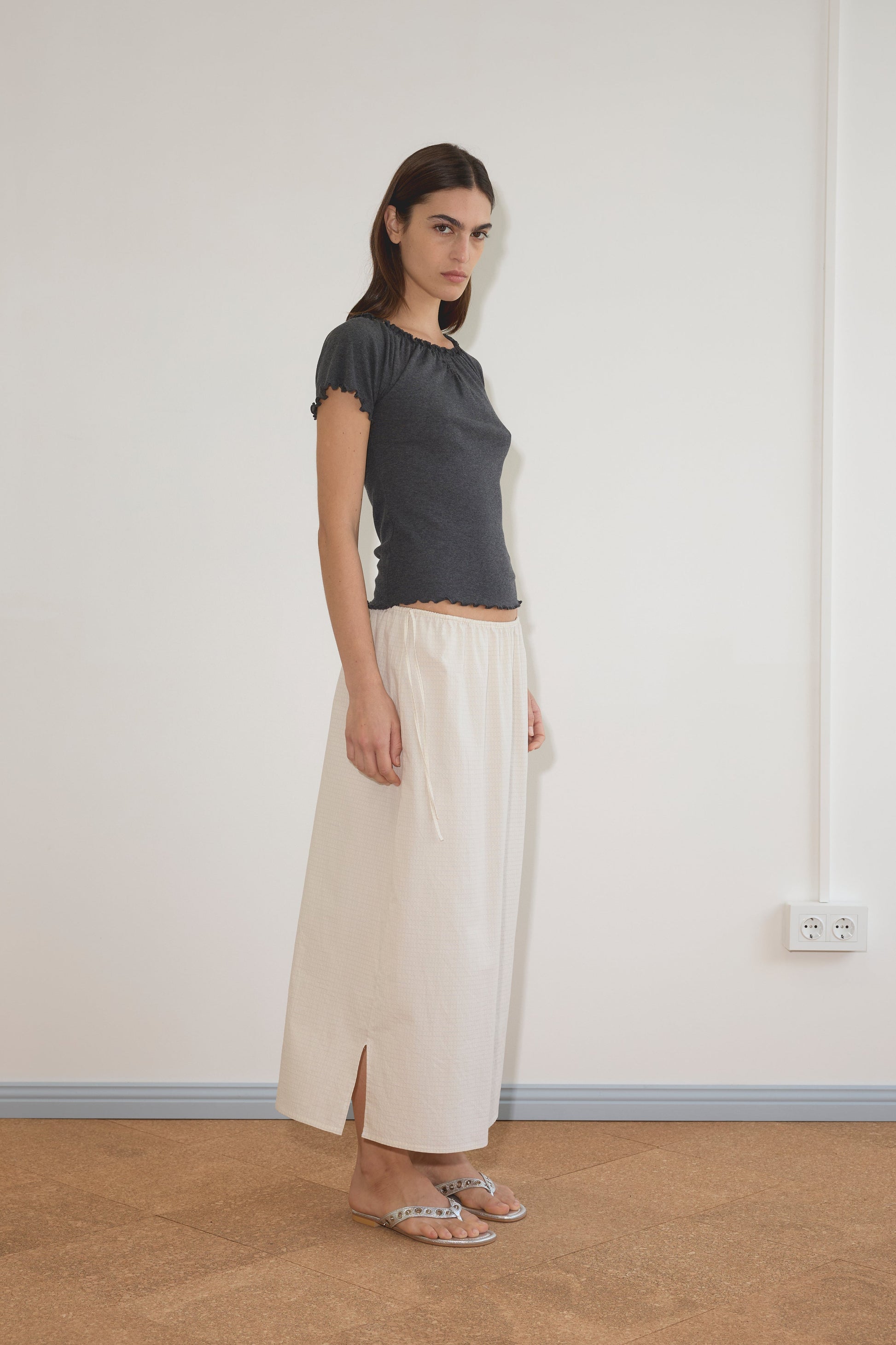 A woman with straight dark hair and a neutral expression stands against a plain wall, wearing the Flounce Tee - Grey by Deiji Studios, a long cream skirt with side slits, and flat sandals.