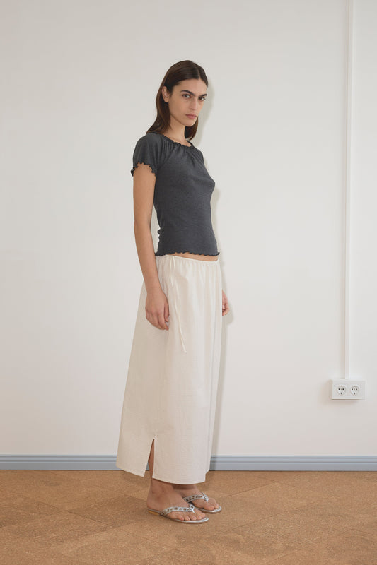 A woman with straight dark hair and a neutral expression stands against a plain wall, wearing the Flounce Tee - Grey by Deiji Studios, a long cream skirt with side slits, and flat sandals.