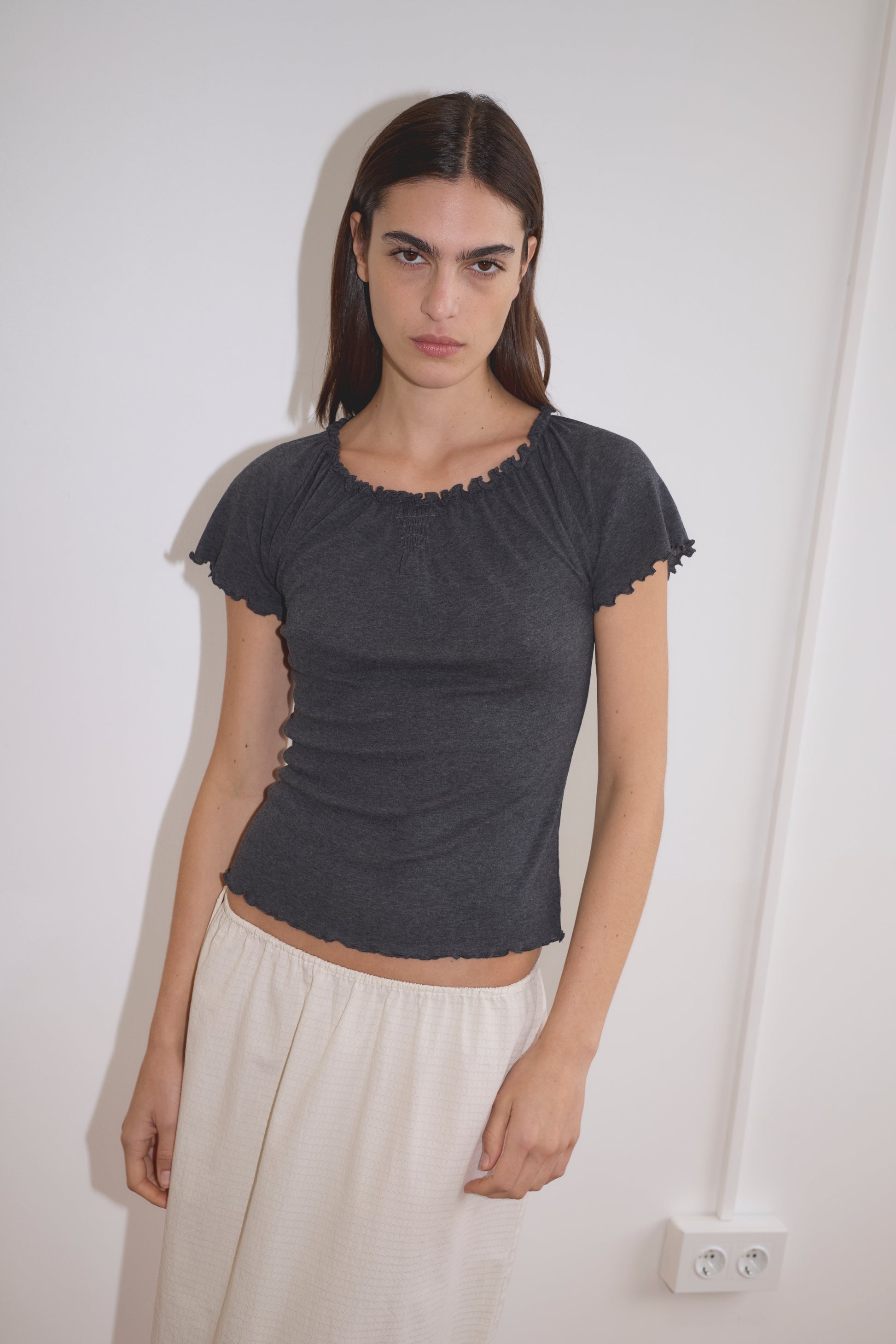 A woman with straight dark hair wears the Flounce Tee - Grey, paired with a light beige skirt, standing indoors against a white wall with an electrical outlet visible.