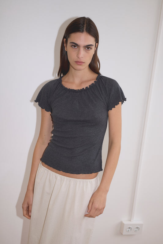 A woman with straight dark hair wears the Flounce Tee - Grey, paired with a light beige skirt, standing indoors against a white wall with an electrical outlet visible.