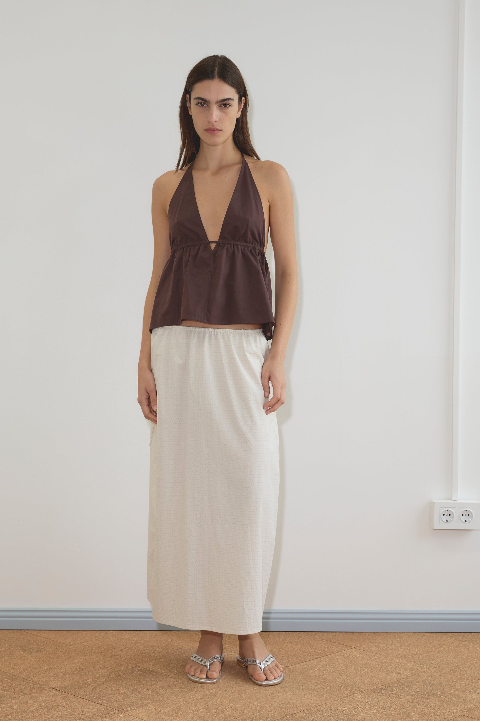 A woman with straight brown hair wears the Button Halter - Umber, a sleeveless brown organic cotton halter top with a deep V-neck, and pairs it with a long white skirt and silver sandals, standing against a plain white wall indoors.