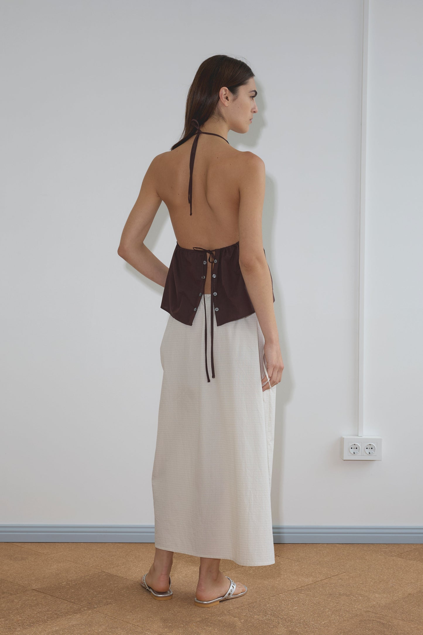 A woman stands indoors facing away, wearing the Button Halter - Umber from Deiji Studios with lace-up details and a long, light beige skirt. She has straight brown hair and is wearing flat sandals.