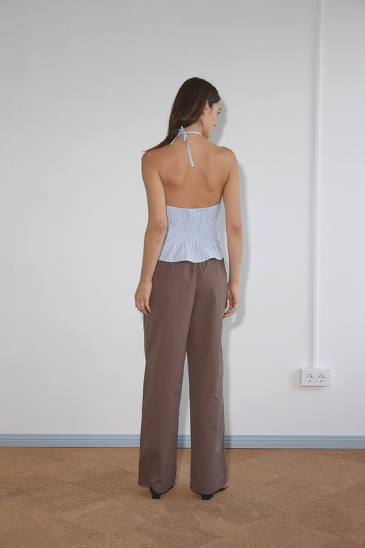 A woman stands facing a white wall, wearing the Strap Top - Mineral Stripe and loose brown pants. Her straight hair is worn down, and she is photographed from behind. A double electrical outlet appears on the wall to her right.