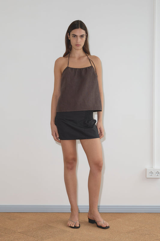 A woman with long dark hair stands indoors against a white wall, wearing the O Ring Halter - Bark top from Deiji Studios, a short black skirt, and black sandals. Her expression is neutral, and her arms are relaxed at her sides.
