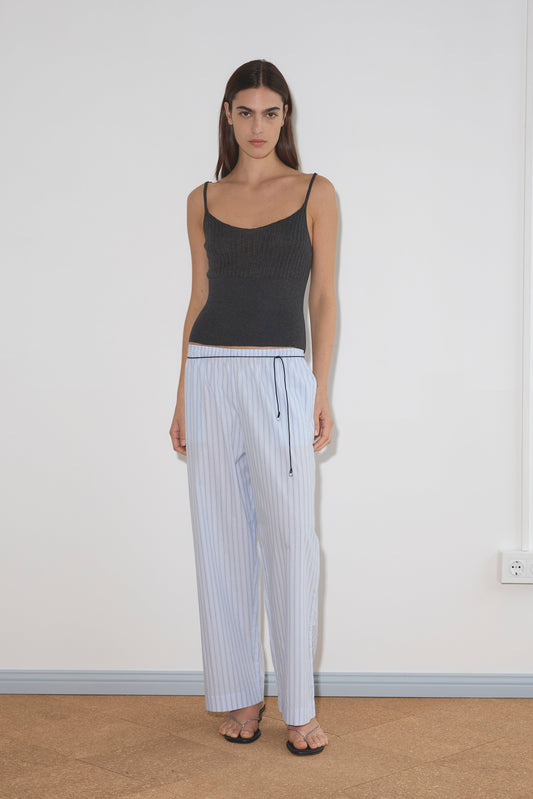 A woman with long brown hair wears the Ease Trouser in light blue with a drawstring, a dark gray spaghetti strap tank top, and black sandals, standing against a plain white wall on a tan floor.