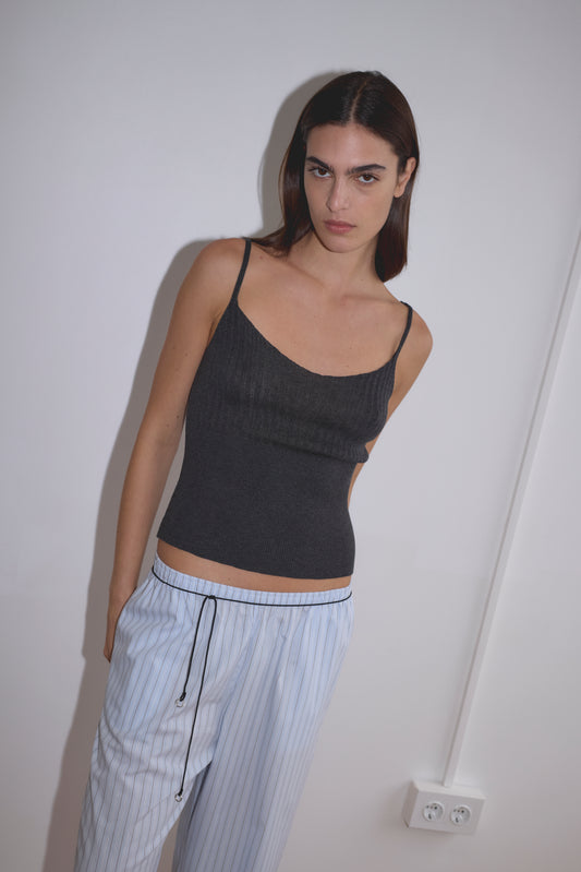 A woman with straight dark hair wears a dark gray sleeveless top, light blue striped pajama pants, and the Multi Ring Belt - Black, standing against a white wall with her hands in her pockets. An electrical outlet is visible near her leg.