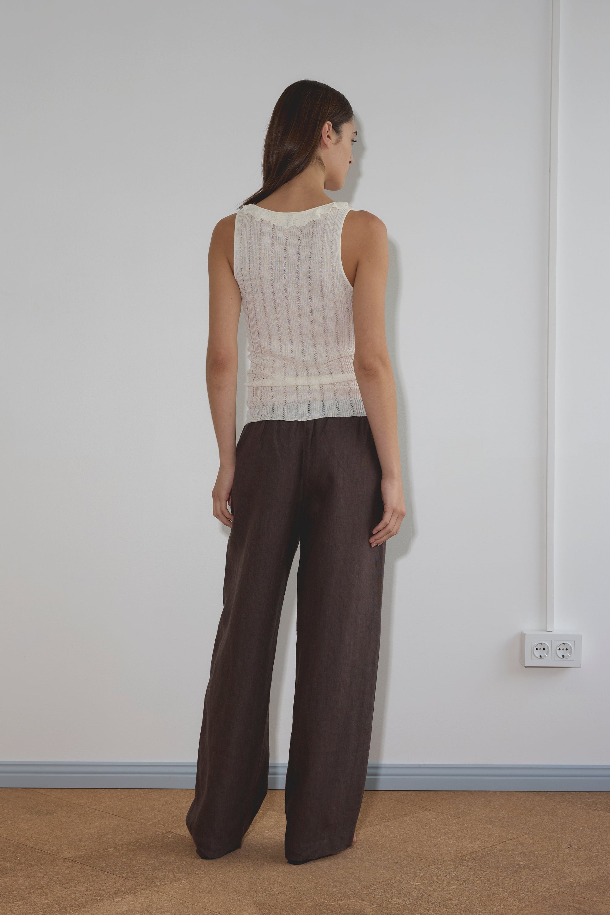 A woman with straight brown hair faces a white wall, wearing the Neck Ruffle Knit - White and loose dark brown pants. The room features a light brown floor and a white electrical outlet on the wall.