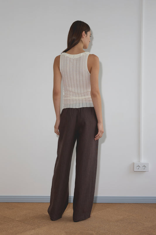 A woman with straight brown hair faces a white wall, wearing the Neck Ruffle Knit - White and loose dark brown pants. The room features a light brown floor and a white electrical outlet on the wall.