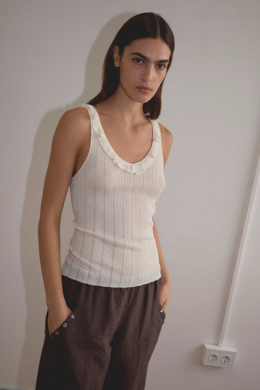 A woman with long dark hair wears the Neck Ruffle Knit - White top and loose dark brown pants, standing against a plain light-colored wall with her hands in her pockets.