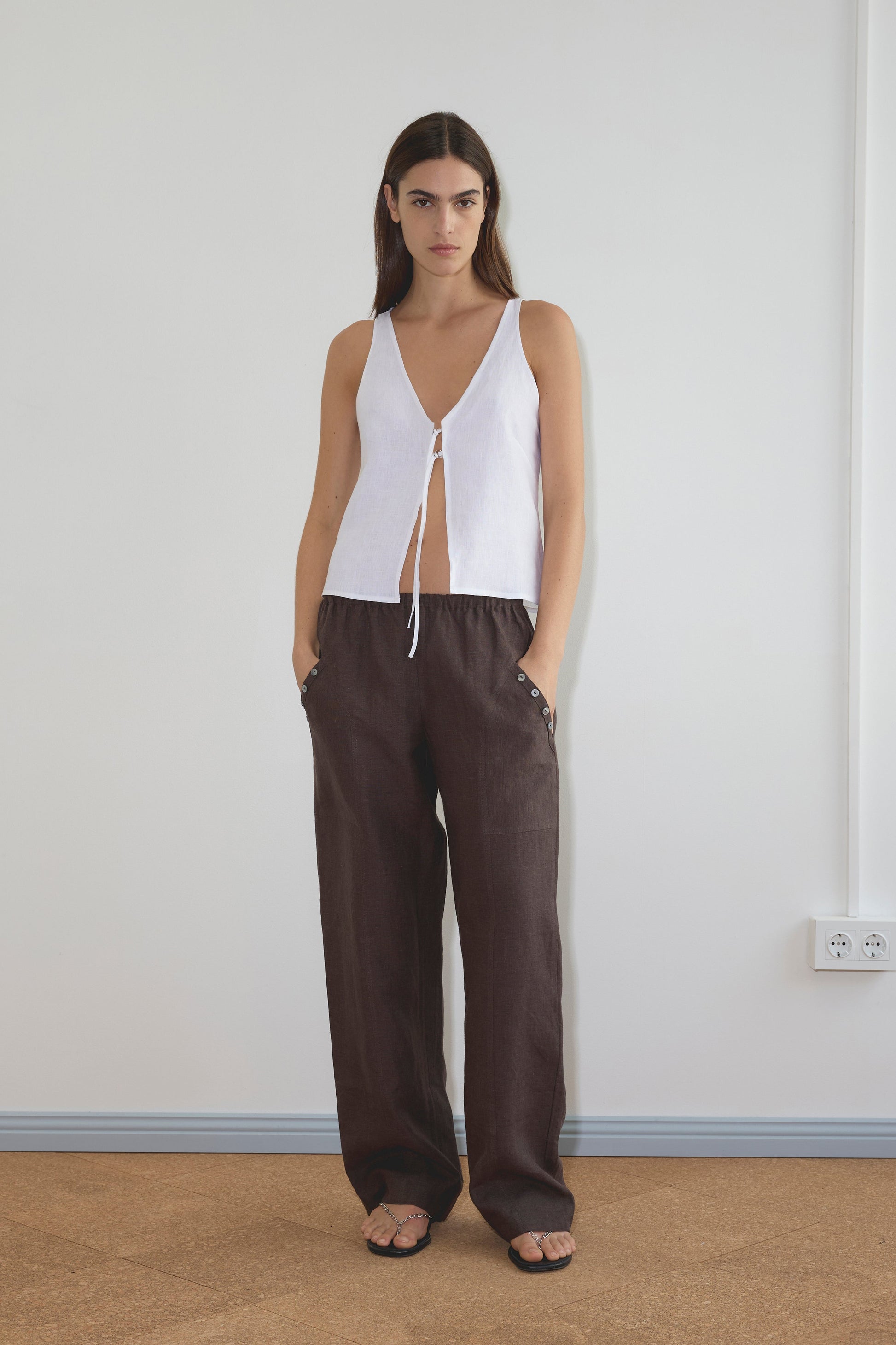 A model wears the Two Part Top - White by Deiji Studios, a sleeveless design with a deep V-neckline, paired with dark brown wide-leg linen trousers with button details and black strappy sandals.
