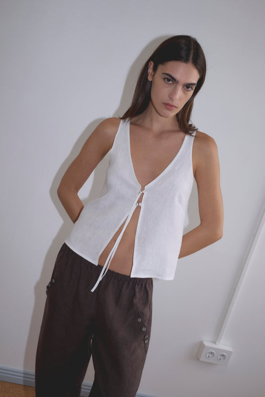 A woman with straight dark hair stands against a white wall, wearing the sleeveless Deiji Studios Two Part Top in white with a deep v-neckline and brown loose-fitting pants. Her hands are in her pockets, and she has a neutral expression.
