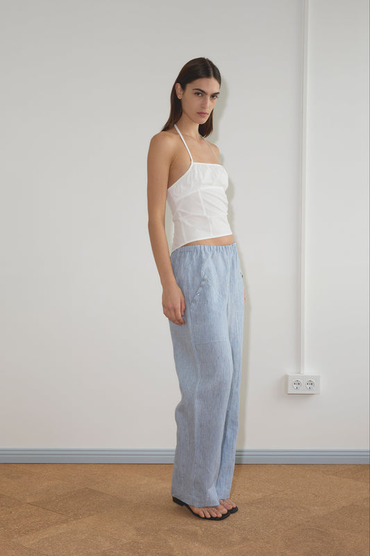 A woman stands indoors barefoot on a tan floor near a white wall, wearing a white sleeveless top with thin straps and the Angle Pocket Pant - Blue Stripe. An electrical outlet is visible in the background.