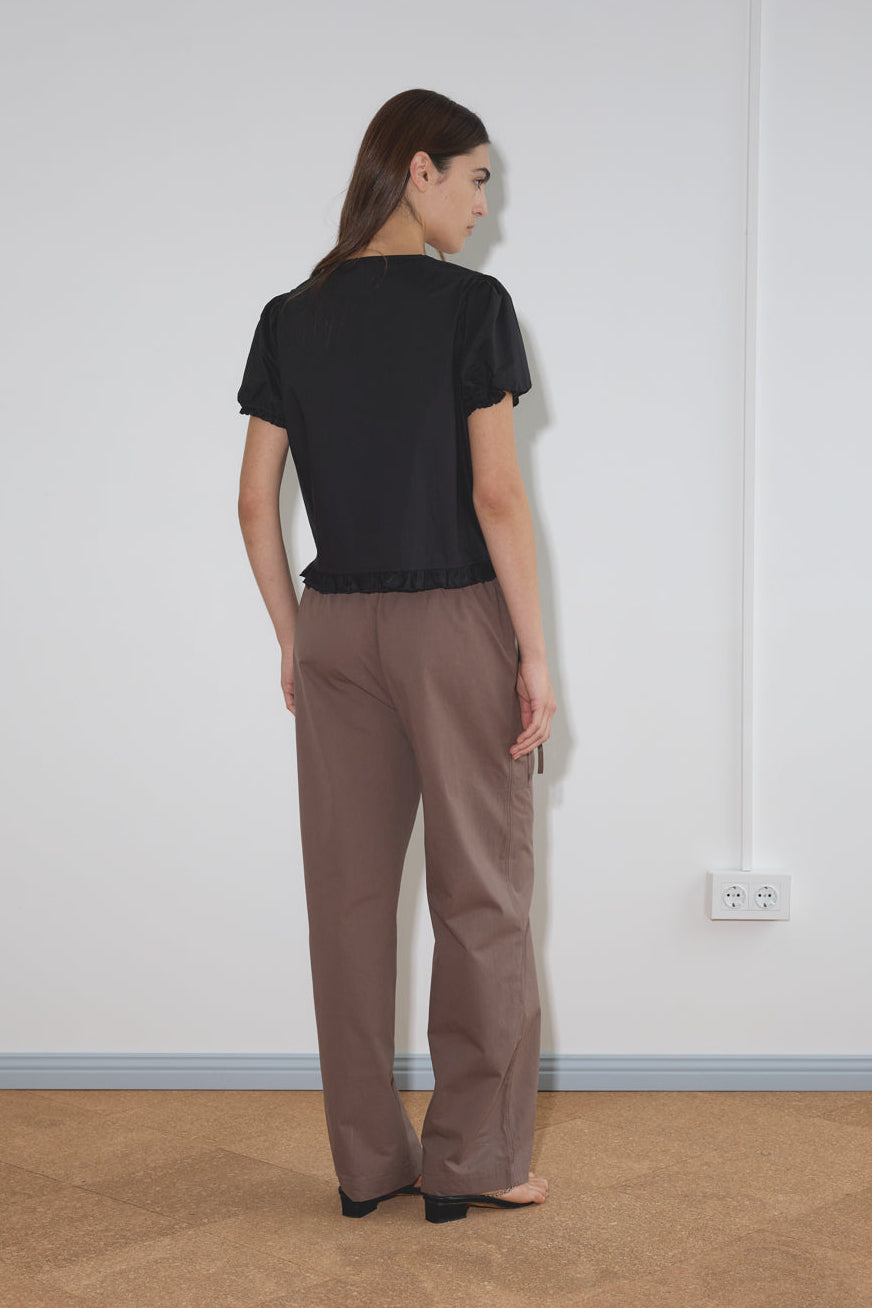 A woman with long brown hair stands facing a white wall, wearing a black Pintuck Top by Deiji Studios with loose brown pants. The photo is taken from behind, showing her back and her left hand resting by her side.