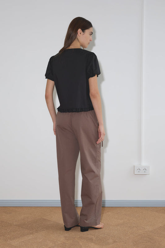 A woman with long brown hair stands facing a white wall, wearing a black Pintuck Top by Deiji Studios with loose brown pants. The photo is taken from behind, showing her back and her left hand resting by her side.