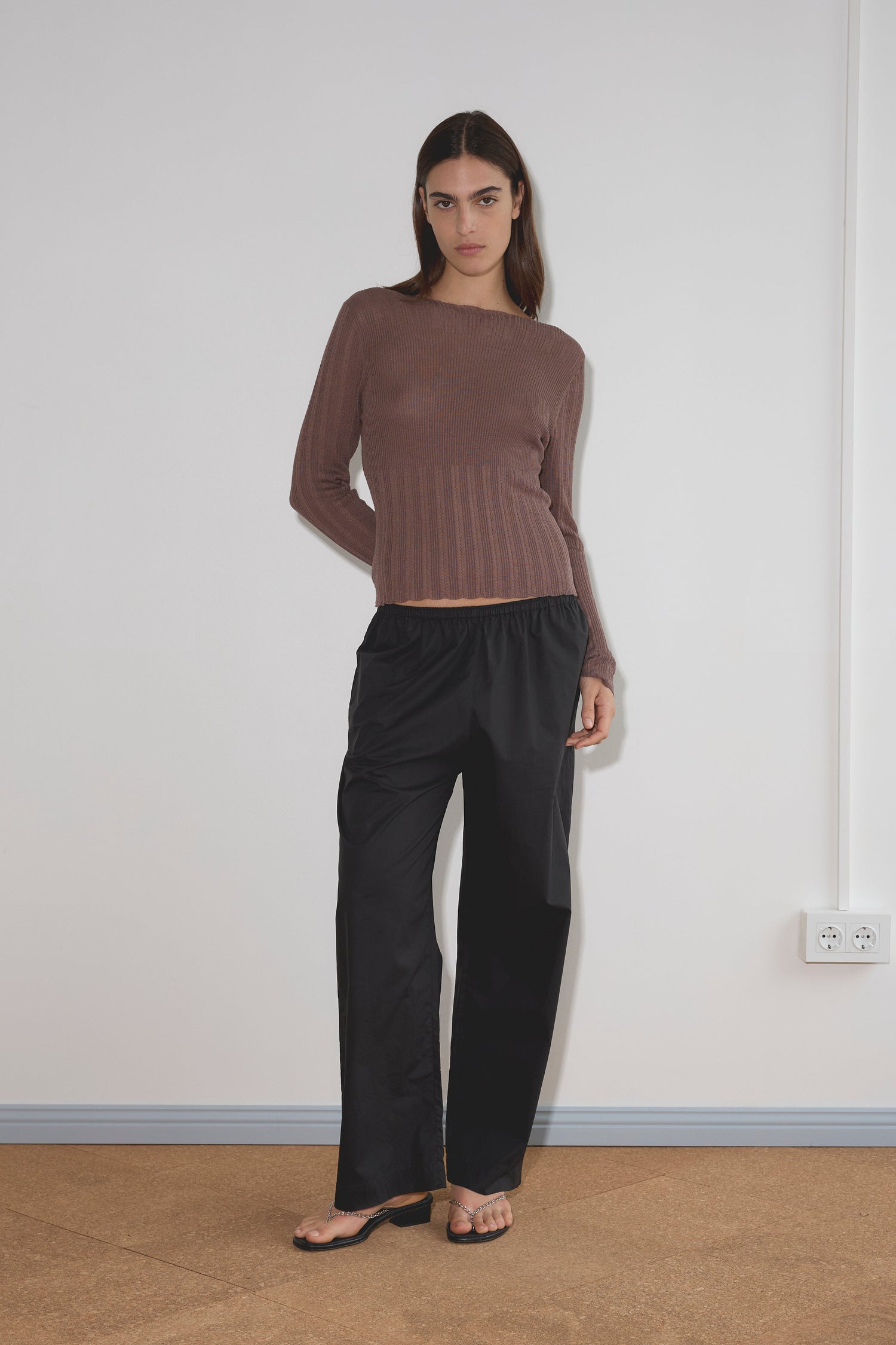 A woman with long dark hair and a neutral expression stands indoors against a white wall, wearing loose black Ease Trousers, a long-sleeve brown ribbed sweater, and black sandals.