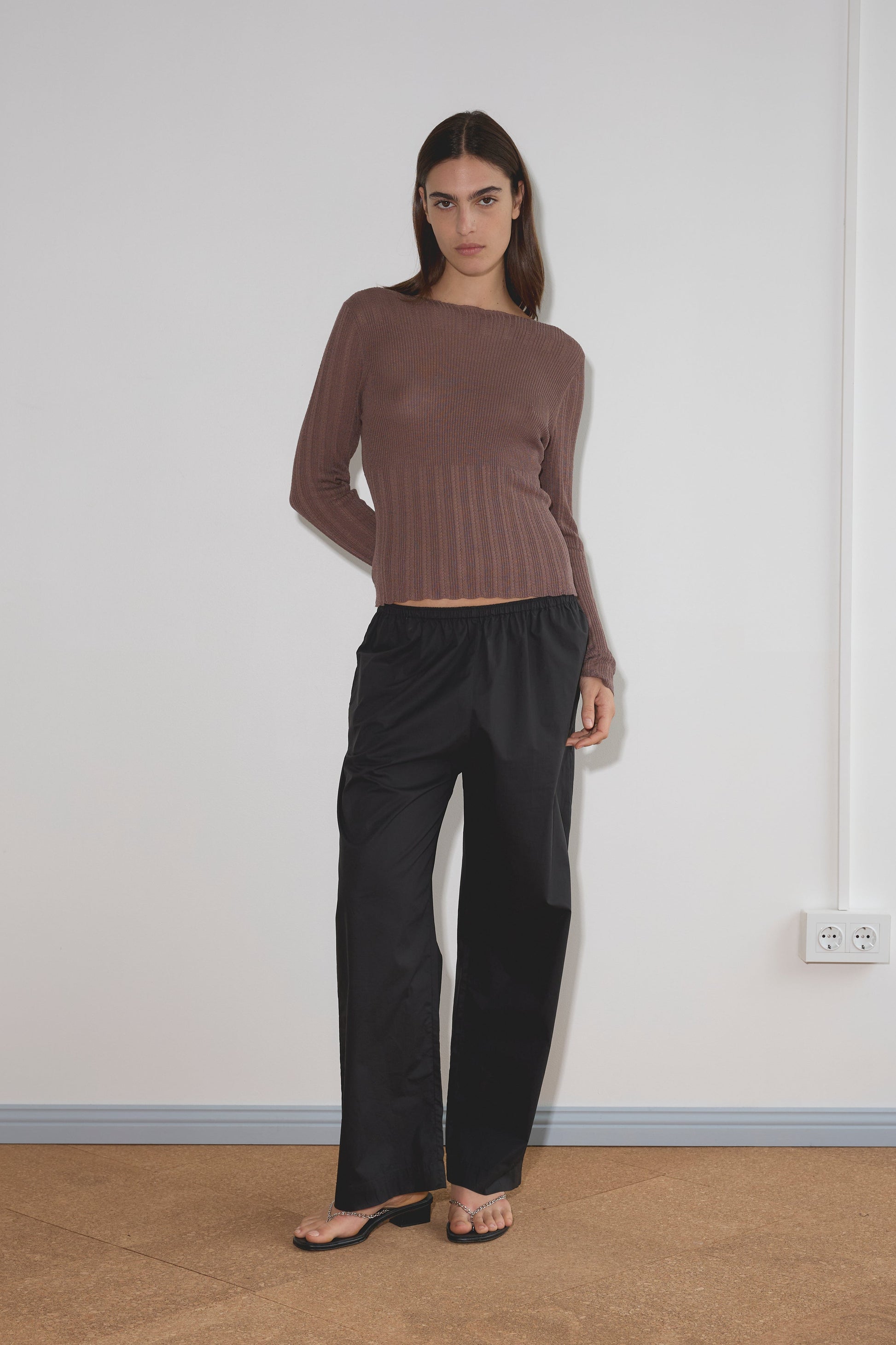 A woman stands indoors by a white wall, wearing a long-sleeved brown ribbed sweater, the Ease Trouser - Black by Deiji Studios in organic cotton, and black sandals. She has straight brown hair and a neutral expression.