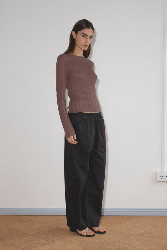 A woman with straight brown hair stands indoors against a white wall, wearing a fitted brown long-sleeve top, black Ease Trouser from Deiji Studios, sandals, and a neutral expression.