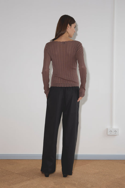A woman with straight brown hair, wearing a long-sleeved brown ribbed sweater and the Ease Trouser - Black, stands facing a white wall with a light brown floor and a power outlet on the right.