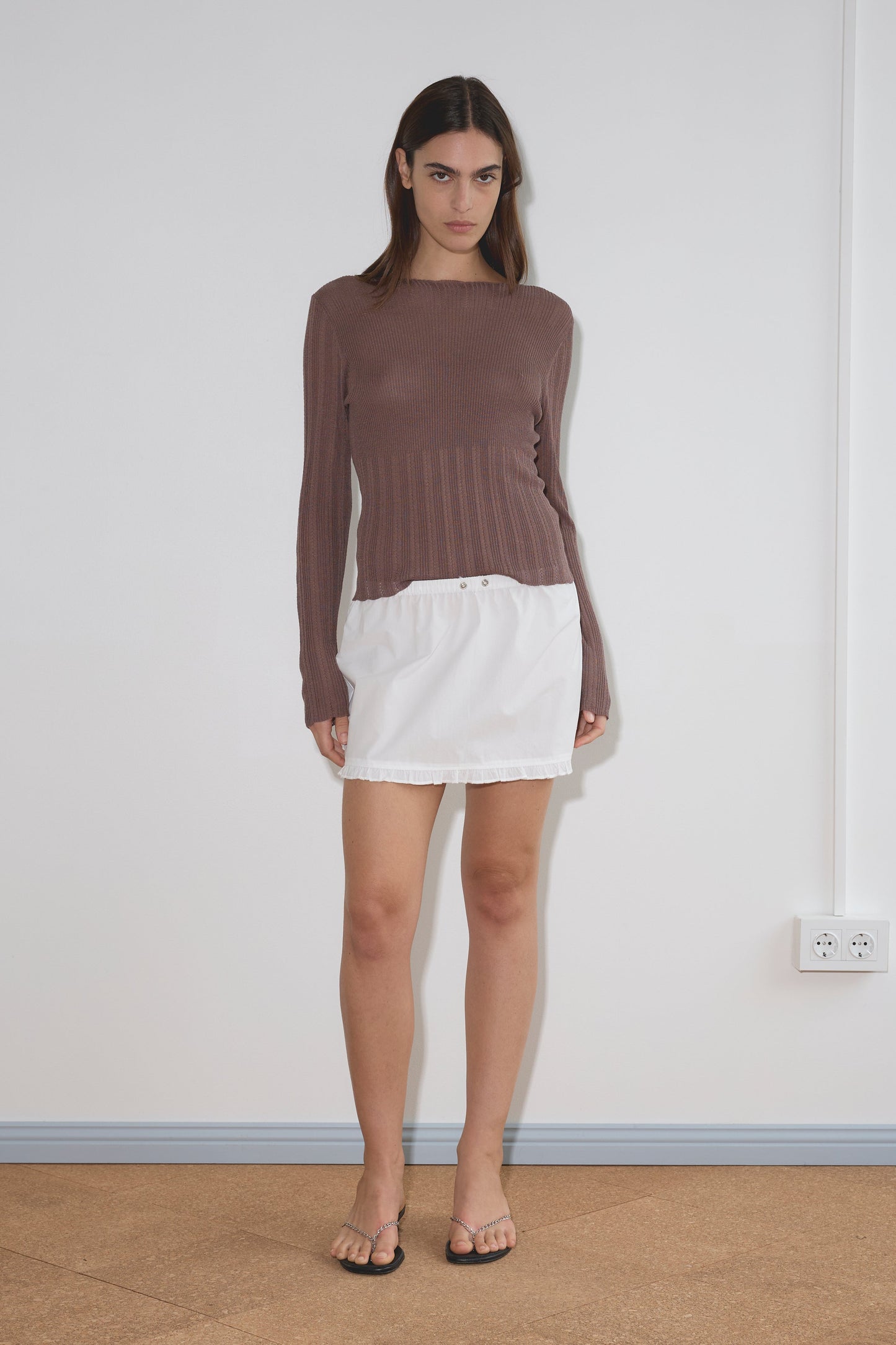 A woman stands indoors against a plain white wall, wearing a long-sleeve brown top, the Flounce Mini - White skirt from Deiji Studios crafted in organic cotton, and black sandals. She has a neutral expression with her arms relaxed by her sides.