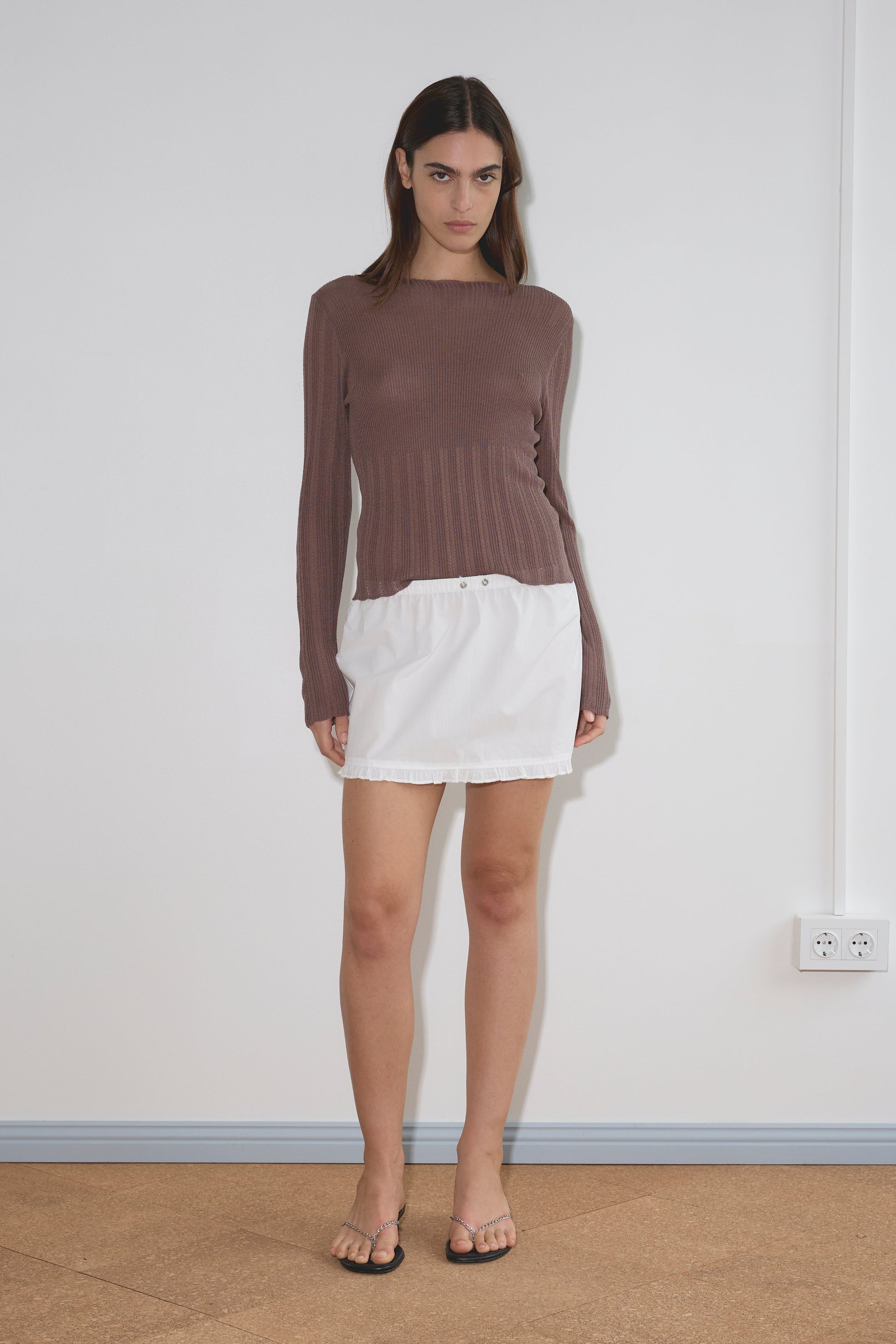 A woman stands indoors against a plain white wall, wearing a long-sleeve brown top, the Flounce Mini - White skirt from Deiji Studios crafted in organic cotton, and black sandals. She has a neutral expression with her arms relaxed by her sides.