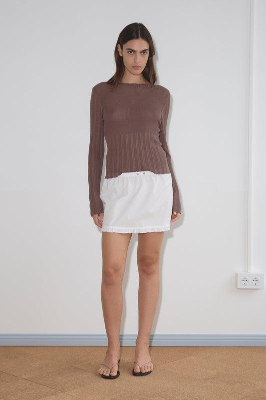 A woman stands indoors against a plain white wall, wearing a long-sleeve brown top, the Flounce Mini - White skirt from Deiji Studios crafted in organic cotton, and black sandals. She has a neutral expression with her arms relaxed by her sides.