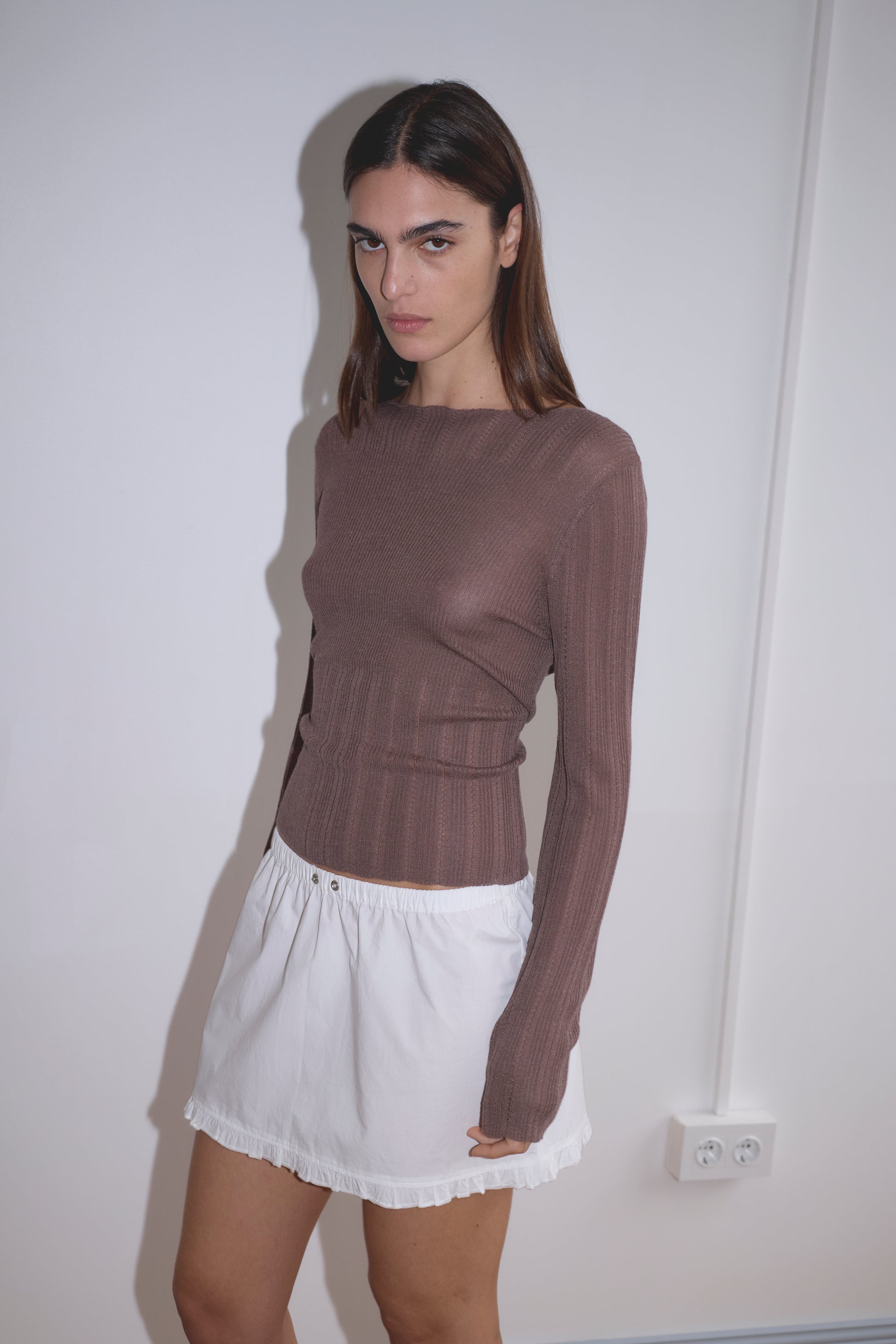 A person with long brown hair stands against a white wall, wearing a fitted brown top and the Flounce Mini - White skirt by Deiji Studios. The look is minimal and neutral, with an electrical outlet visible in the background.