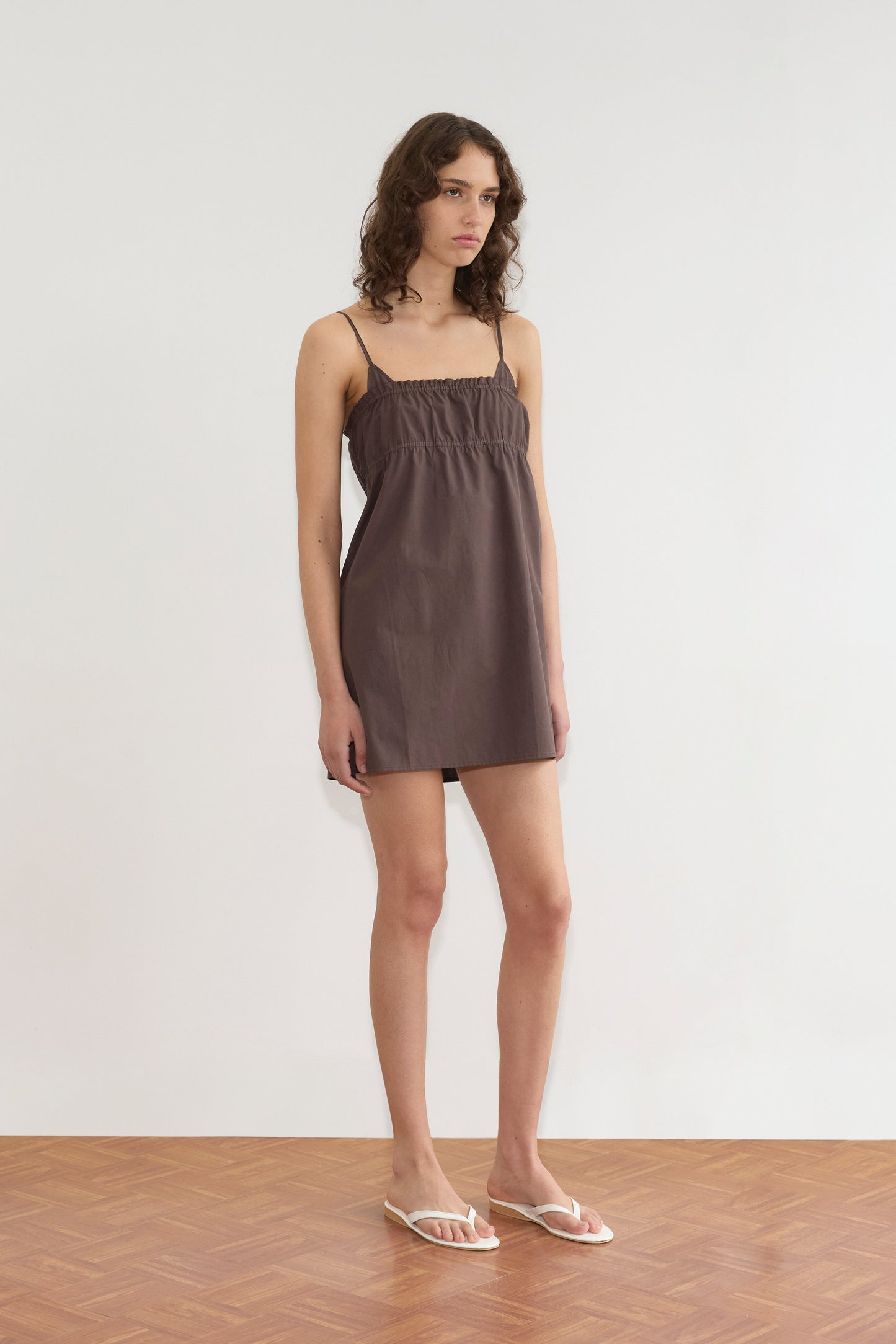 A person with wavy brown hair wears the Edge Dress - Sepia from Deiji Studios and white sandals, standing indoors on a wooden floor against a plain white wall.