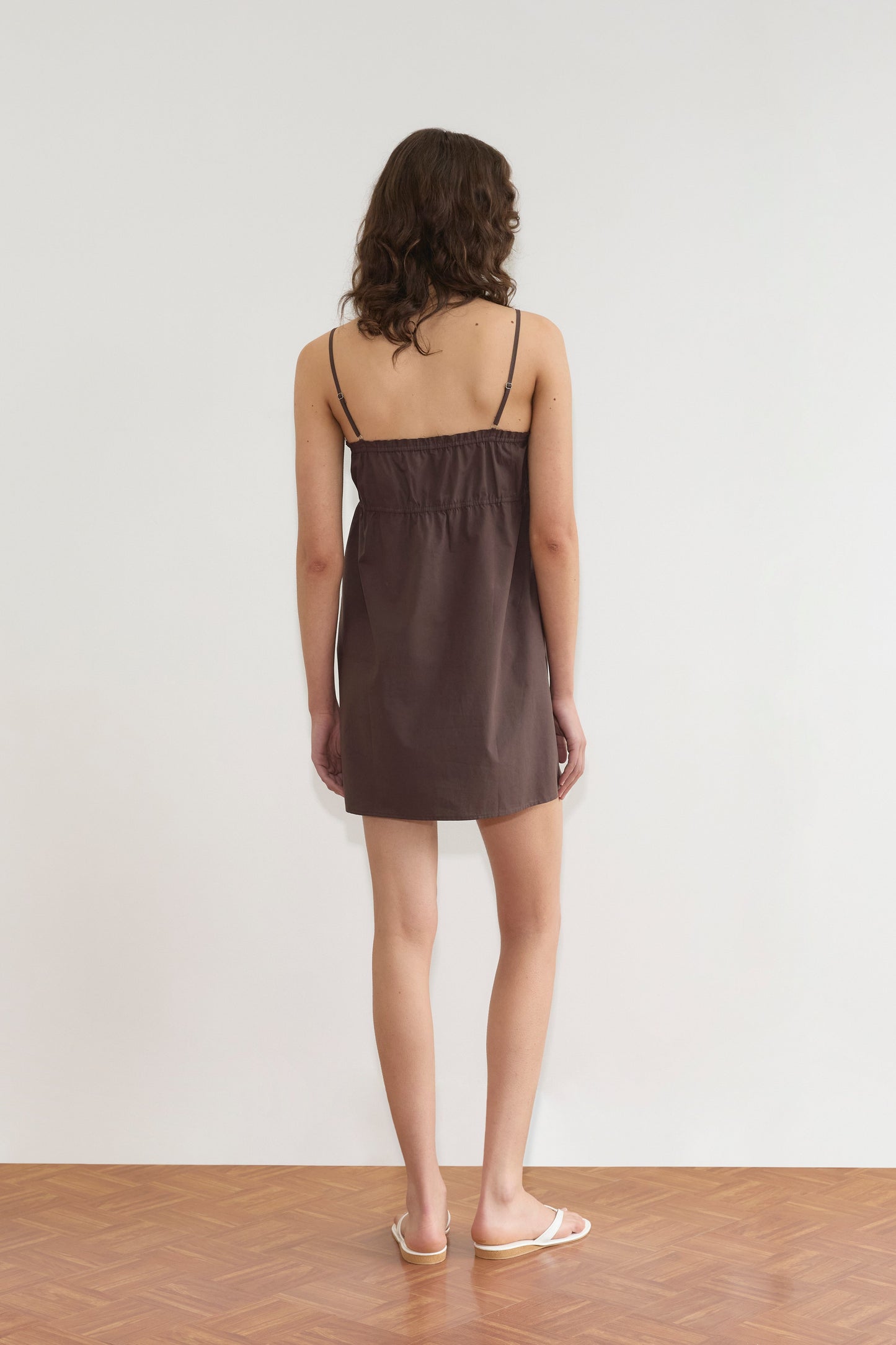 A woman with wavy brown hair stands facing a white wall on a wooden floor, wearing the Deiji Studios Edge Dress in Sepia and white sandals, her arms relaxed by her sides.