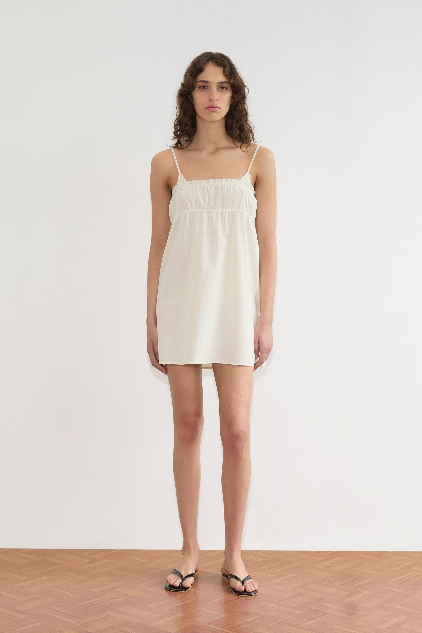 A person with wavy brown hair stands indoors on a wooden floor, wearing the sleeveless Edge Dress - Citrus Stripe and black sandals, facing forward against a plain white background.