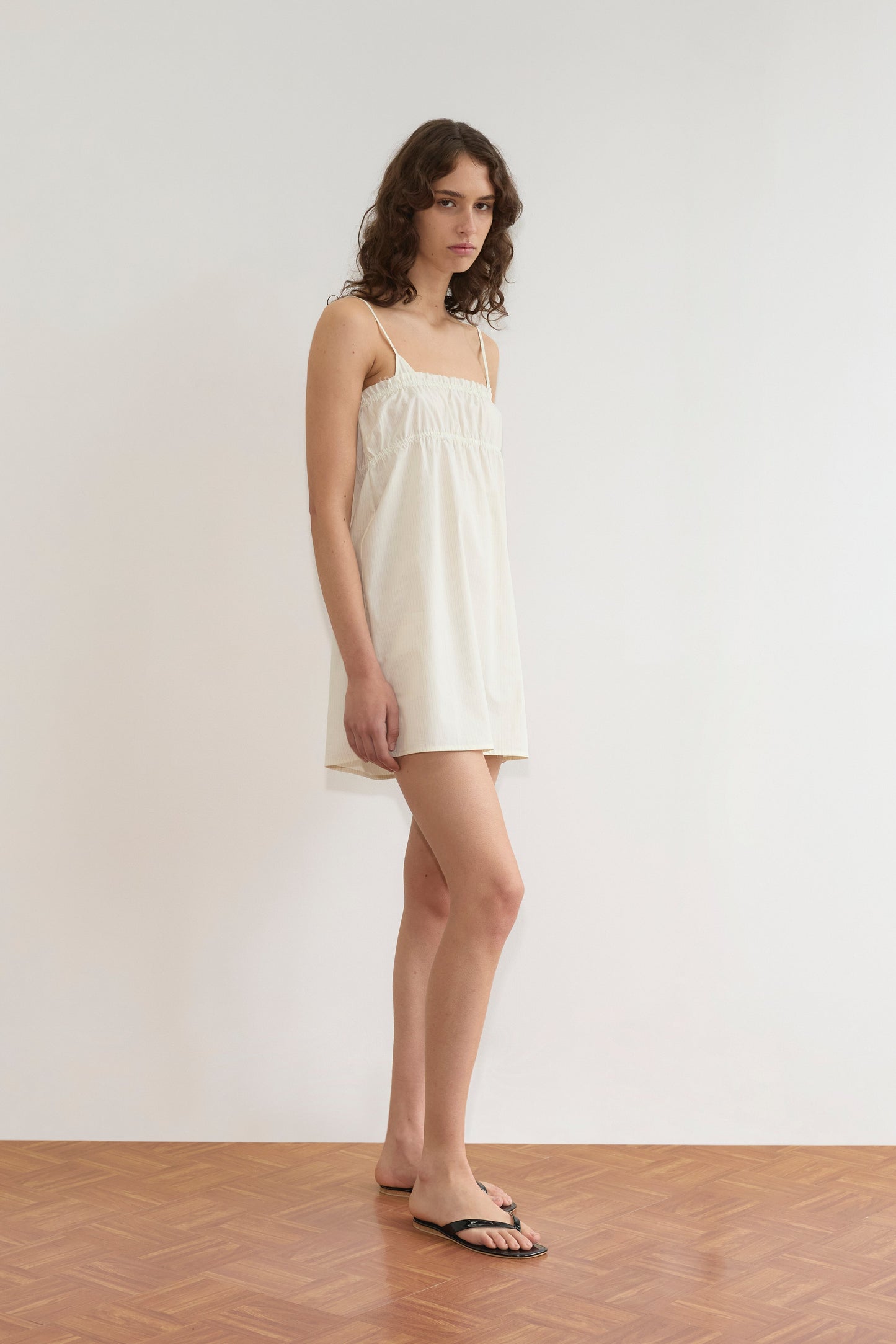 A person with wavy brown hair wearing Deiji Studios’ Edge Dress - Citrus Stripe and black sandals stands on a wooden floor against a plain white wall, looking toward the camera.