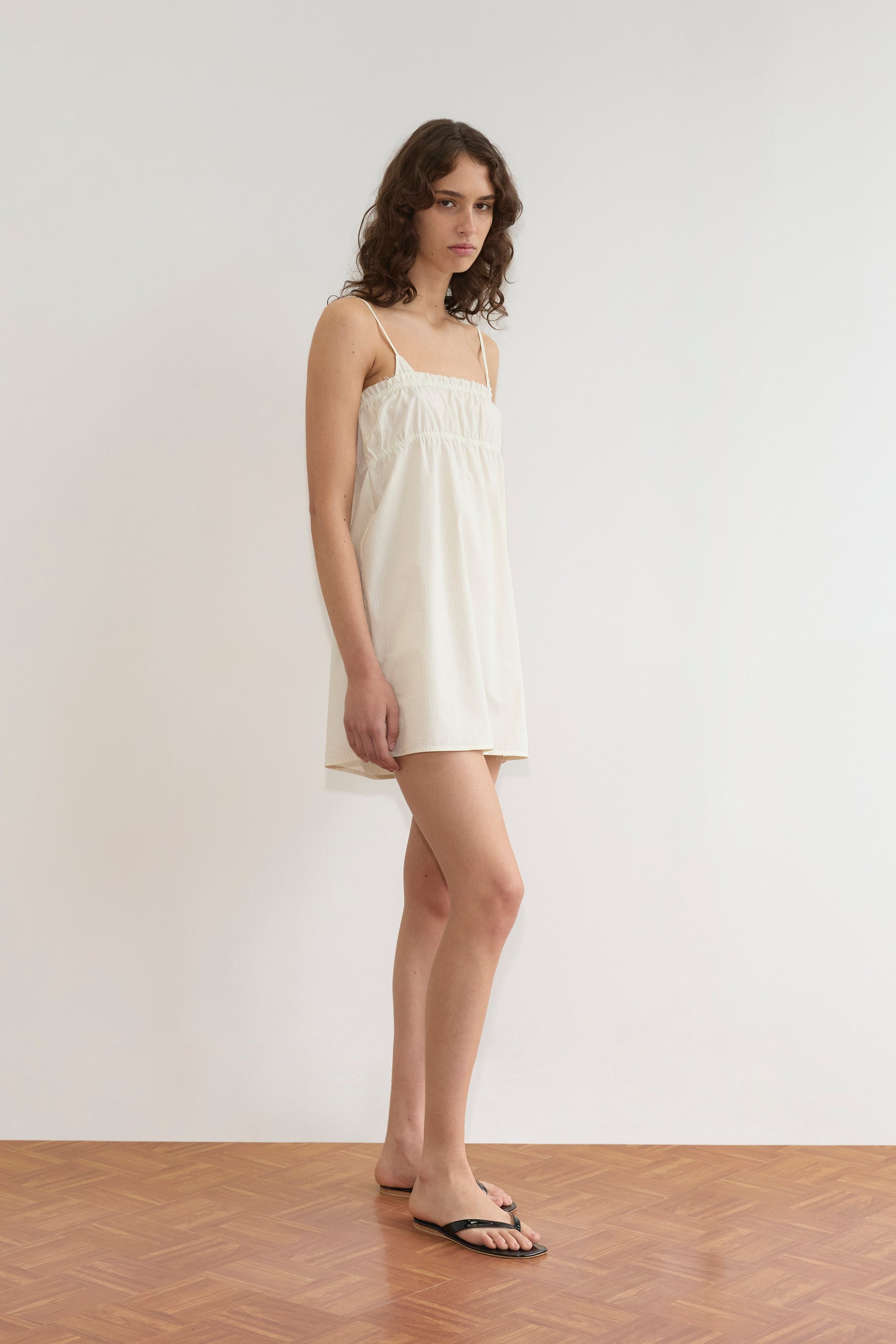 A person with wavy brown hair wearing Deiji Studios’ Edge Dress - Citrus Stripe and black sandals stands on a wooden floor against a plain white wall, looking toward the camera.