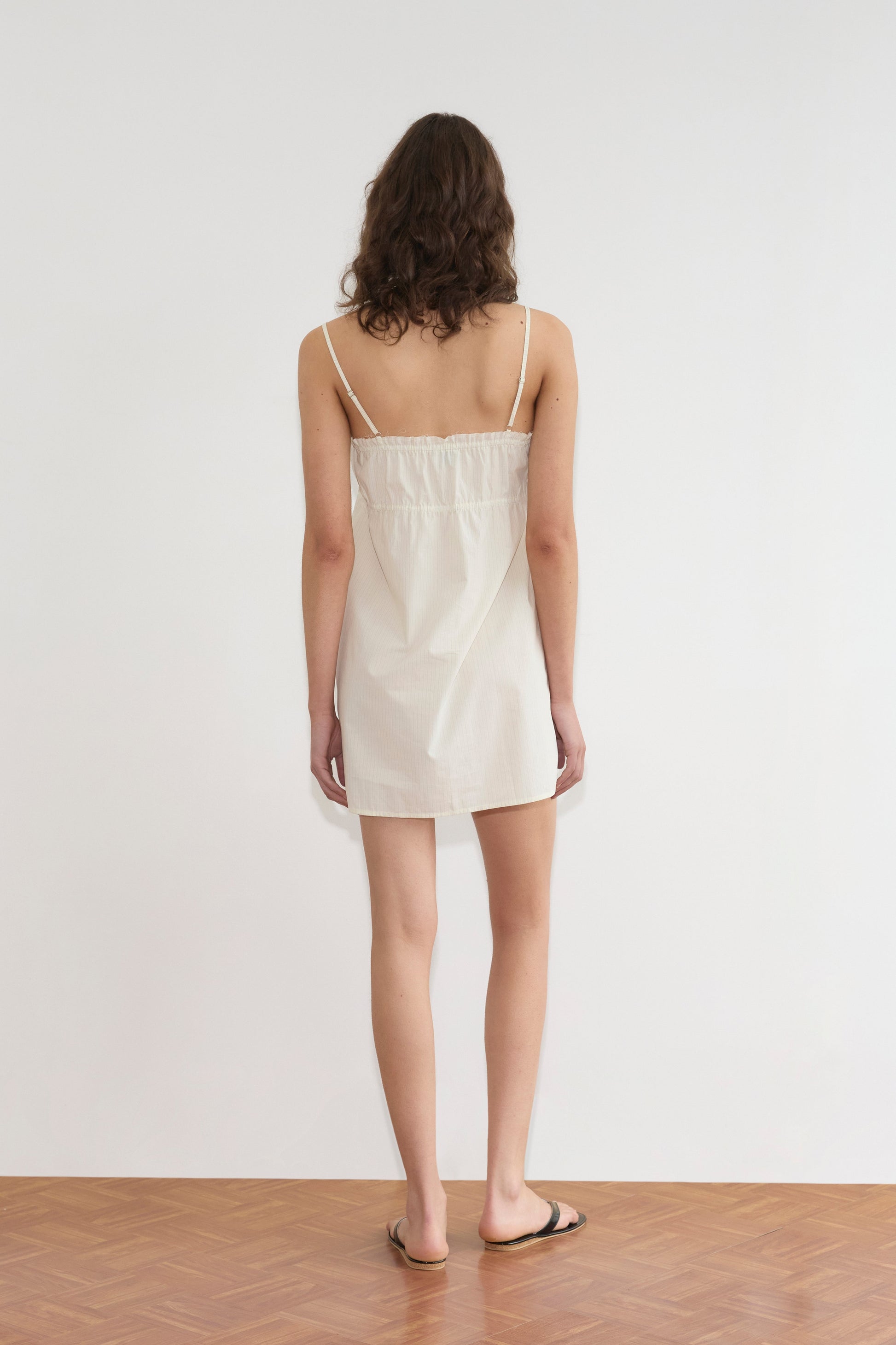 A person with shoulder-length brown hair stands facing a white wall, wearing Deiji Studios' Edge Dress in Citrus Stripe and black slide sandals on a wooden floor.