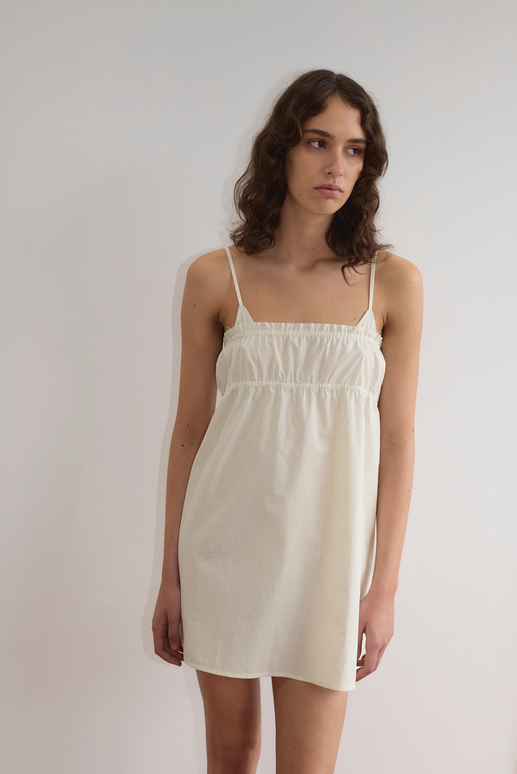 A woman with wavy brown hair wears a sleeveless Edge Dress - Citrus Stripe with thin straps, standing against a plain white wall. She has a neutral expression and her arms are relaxed at her sides.