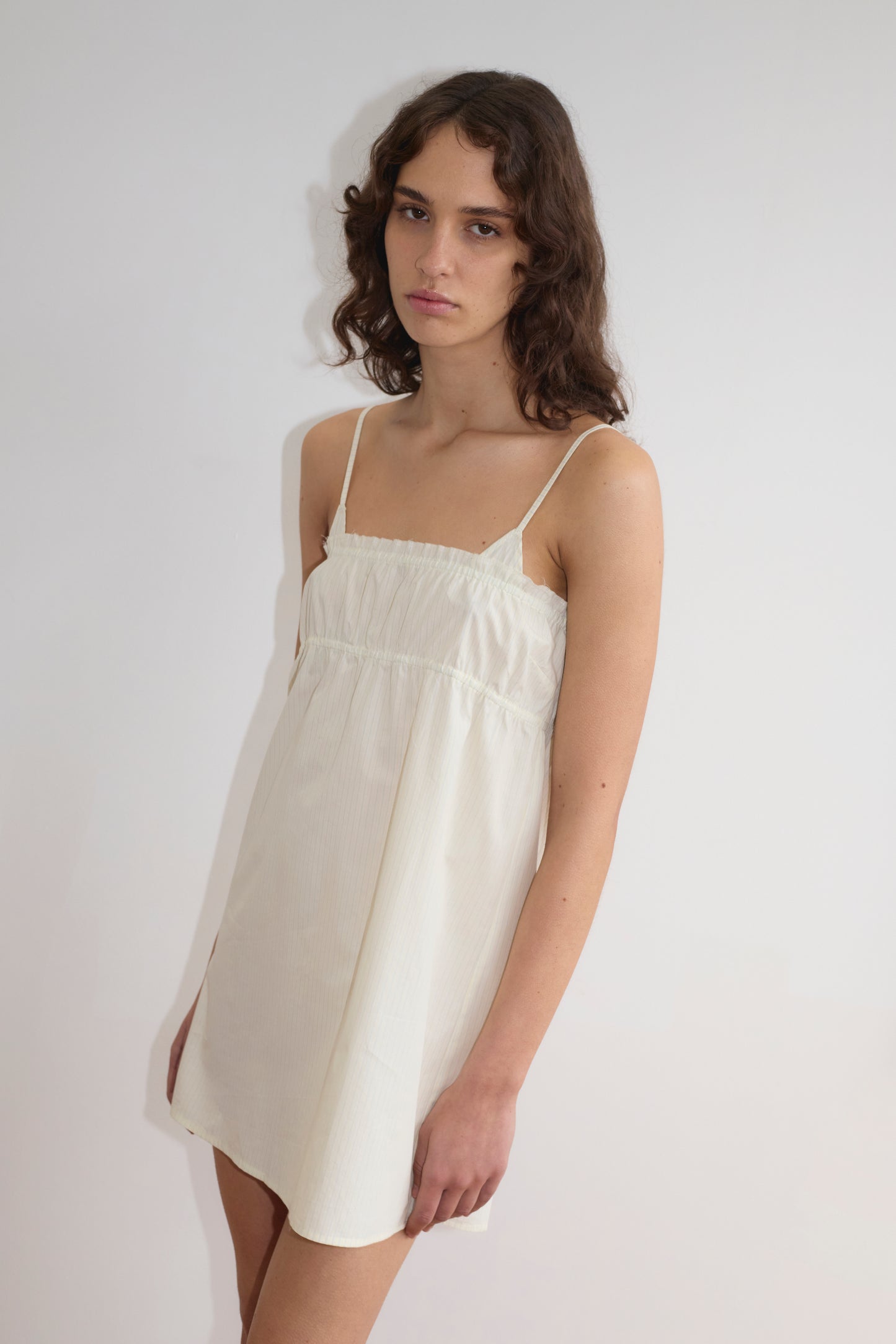 A person with long, wavy brown hair wears a sleeveless, light-colored Deiji Studios Edge Dress in Citrus Stripe and stands against a plain white background, looking at the camera with a neutral expression.