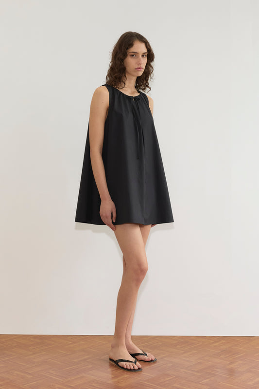 A woman with wavy brown hair stands indoors by a white wall, wearing the Deiji Studios Drawcord Dress - Black with an a-line silhouette and black flip-flops. She gazes to the side with a neutral expression.