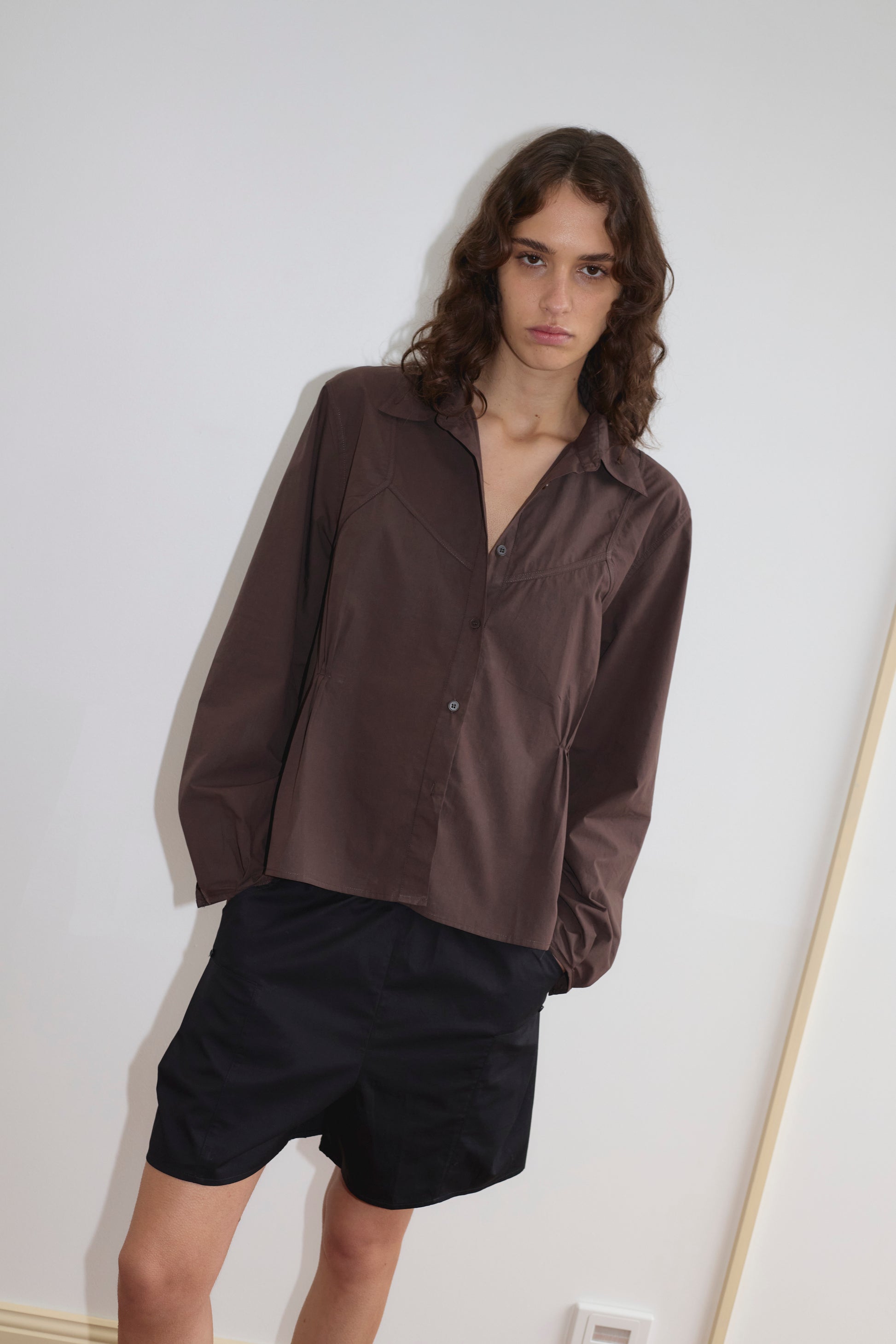 A person with wavy brown hair wears the Deiji Studios Cami Overshirt in Sepia, paired with black shorts, standing against a plain white wall and looking at the camera with a neutral expression.