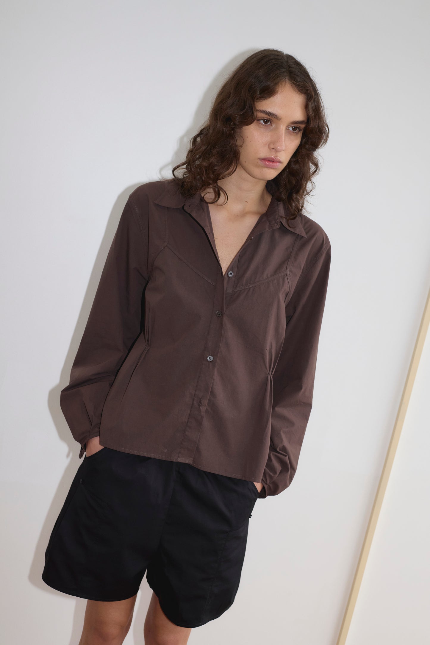 A person with wavy brown hair wears a brown Deiji Studios button-up shirt and relaxed mid-rise Pleat Short in black, standing against a plain white wall with hands in pockets, looking slightly downwards.