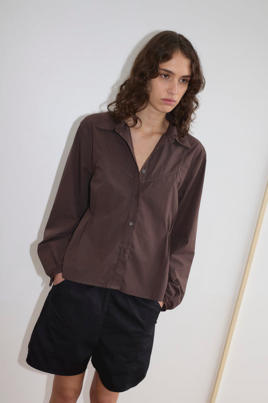 A person with wavy brown hair wears a brown Deiji Studios button-up shirt and relaxed mid-rise Pleat Short in black, standing against a plain white wall with hands in pockets, looking slightly downwards.